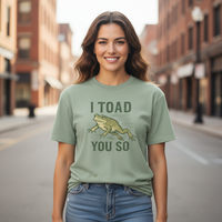 Load image into Gallery viewer, Woman wearing a Bay green t-shirt with a frog graphic and text on a city street.